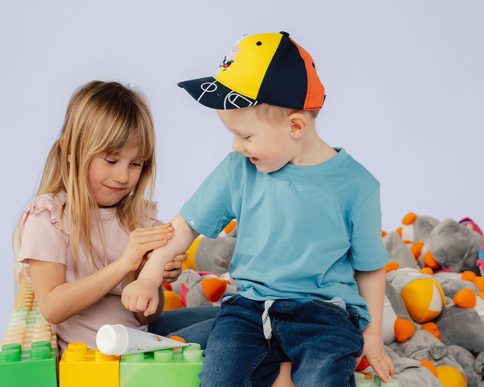 Zwei Kinder spielen mit Bauklötzen, ein Kind cremt dem anderen den Arm ein.