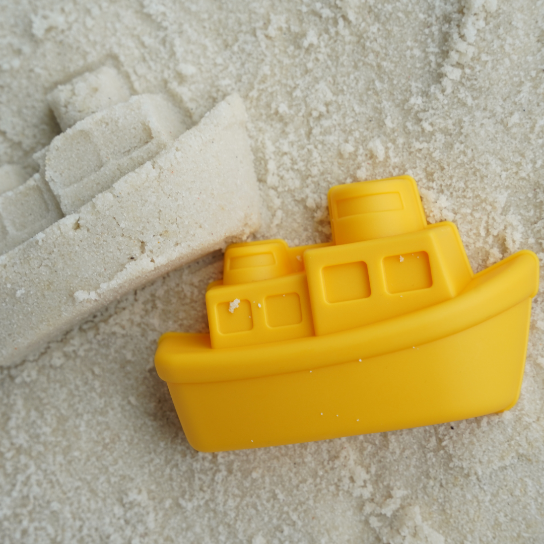 Gelbes Schiff-Sandförmchen im Sand neben dem daraus geformten Schiff.