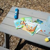 Pauli-Büchlein ausgebreitet auf einem Picknicktisch, daneben Sonnenstick und eine Kappe.