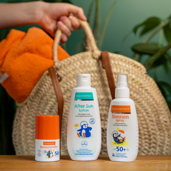 Der PAEDIPROTECT Sonnenroller, Sonnenspray und After Sun Lotion auf einem Tisch mit einer Hand, die eine Strandtasche mit einem Strandtuch darin hält