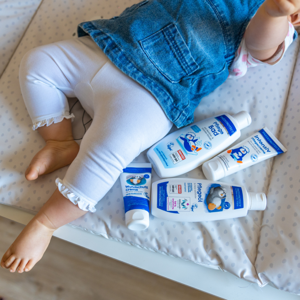 Ein Baby mit einem Jeanskleid und einer weißen Stoffhose liegt auf einer Wickelunterlage mit grauen Punkten. Neben dem Baby befinden sich Pflegeprodukte.