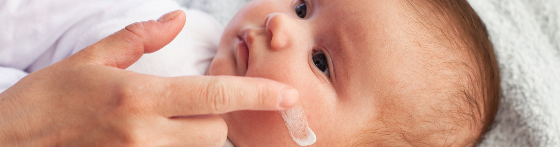 Baby mit Hautreizungen, das sanft mit Gesichtscreme eingecremt wird neugeborenes Baby, das sich hinlegt, während seine Mutter ihm mit dem Finger sanft Gesichtscreme aufträgt