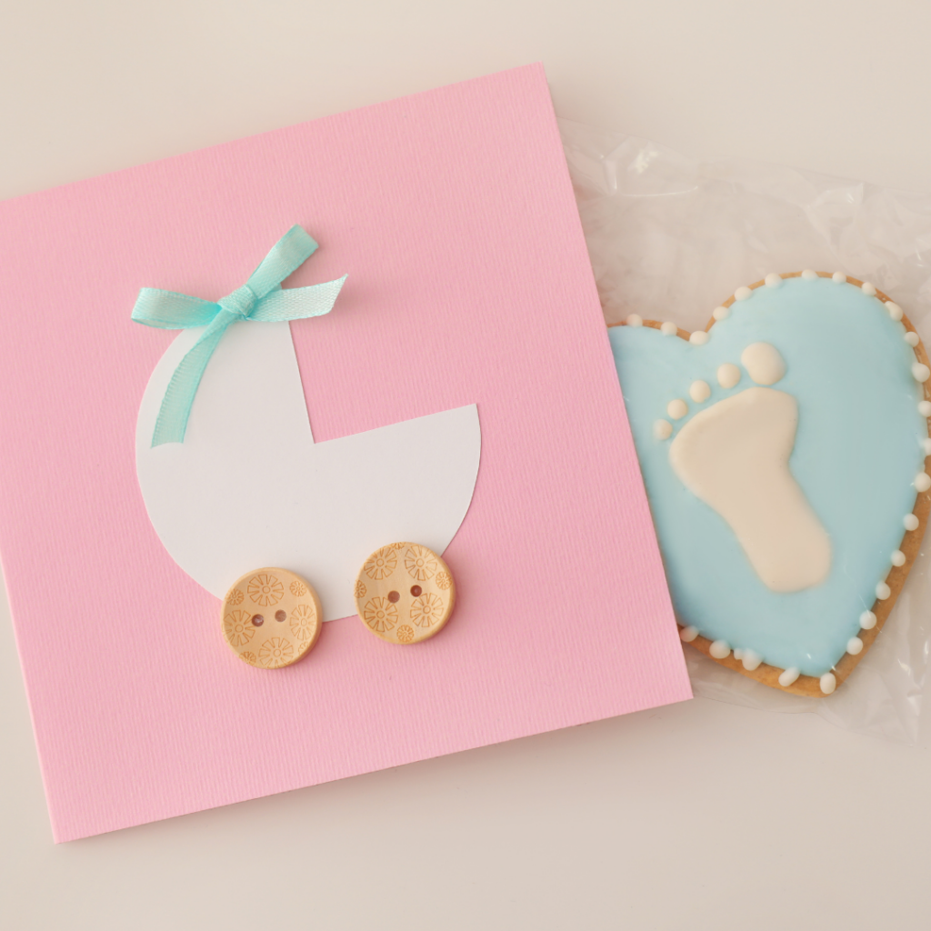 Eine rosa Babykarte mit einem Kinderwagen darauf neben einem blauen Keks mit einem kleinen Fuß darauf