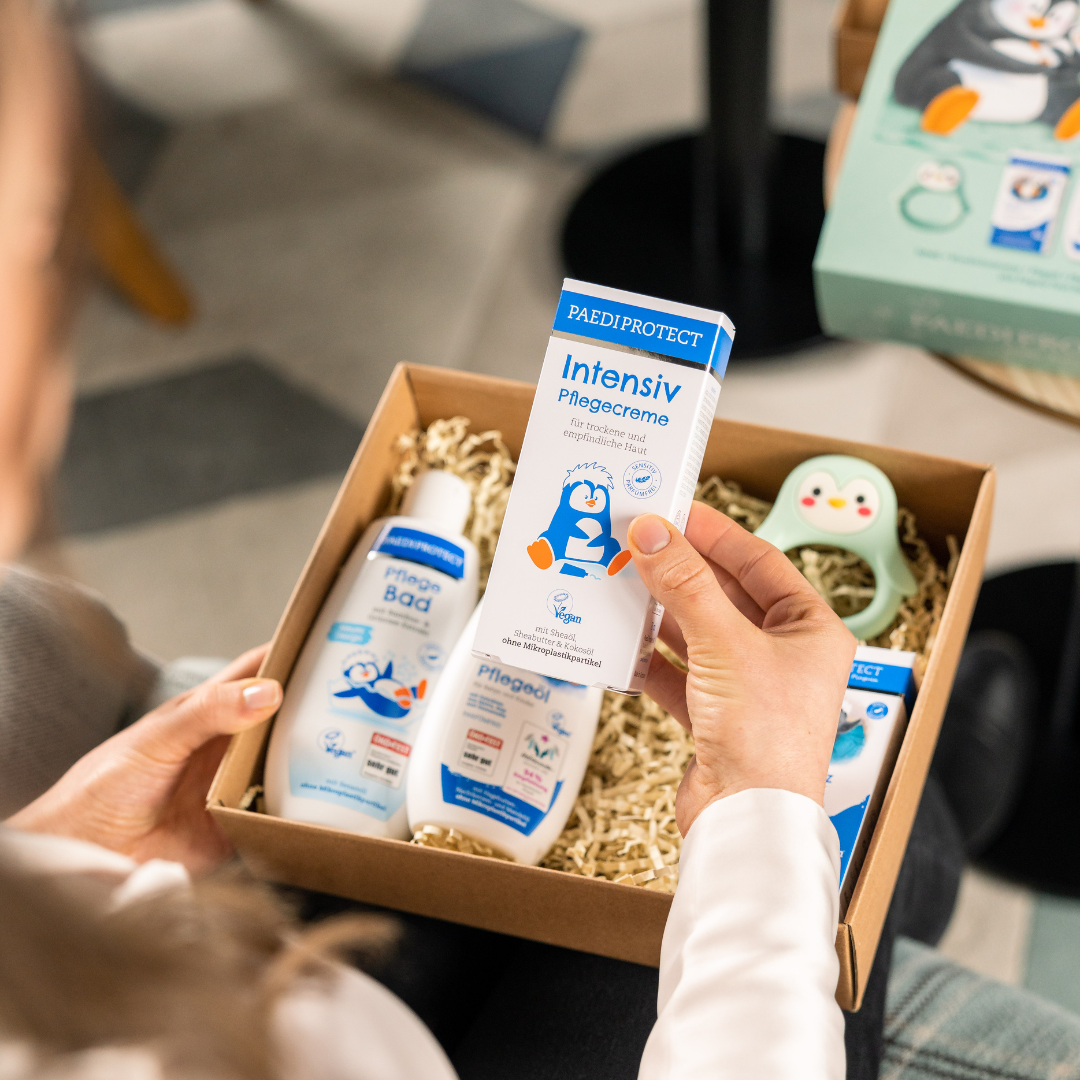 Eine Frau sitzt auf einem Stuhl, hält das geöffnete Hallo Baby Set auf dem Schoß und zeigt die Intensiv Pflegecreme mit der Hand in die Kamera. Die Banderole des Hallo Baby Sets befindet sich im Hintergrund auf einem kleinen Tisch.